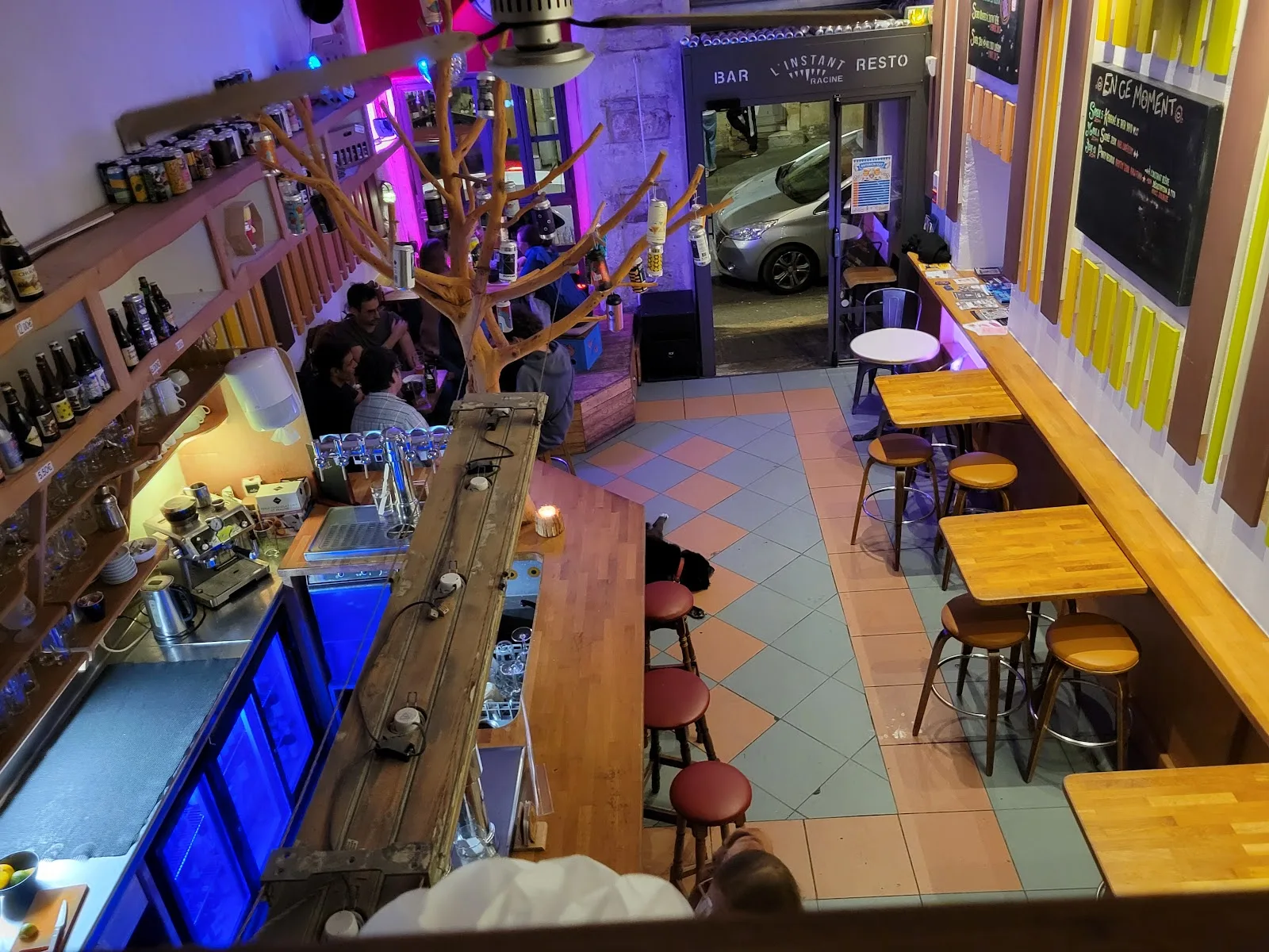 La salle de L'Instant T vue de la mezzanine, le soir : le comptoir, les tireuses, les tables en bois et la porte ouverte sur la rue Racine