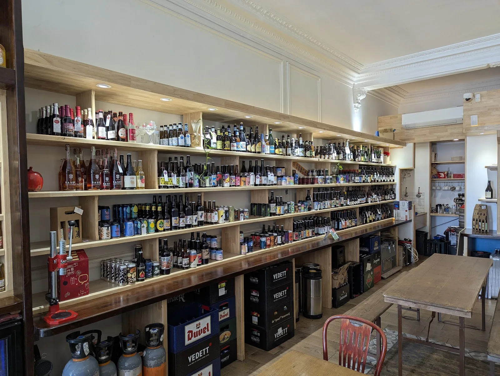 Le mur de la cave de L'Instant T : quatre étagères en bois clair remplies de bouteilles et de canettes de bière, du sol au plafond