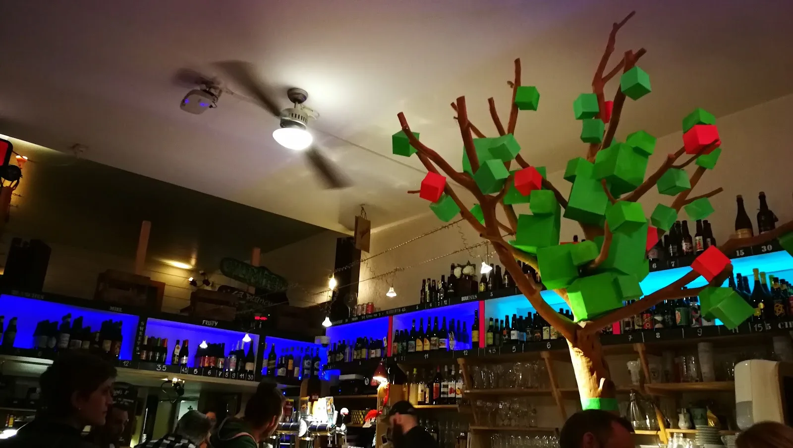 Le comptoir de L'Instant T le soir : l'arbre en bois aux feuilles cubiques vertes et rouges, devant les étagères de bouteilles rétroéclairées en bleu