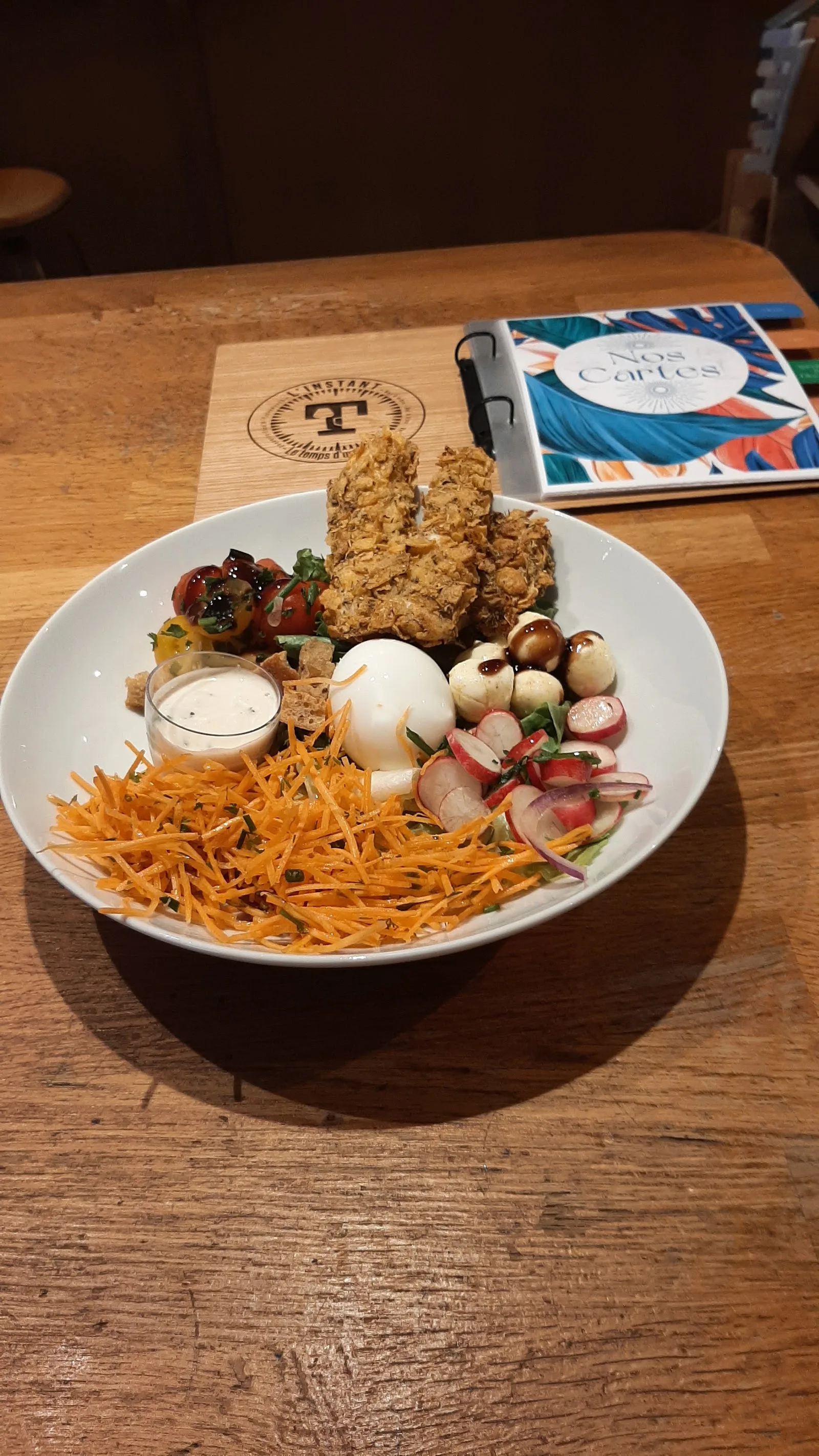 Une assiette servie à L'Instant T : carottes râpées, radis, tomates, œuf, croustillant de poulet, à côté du carnet « Nos Cartes »