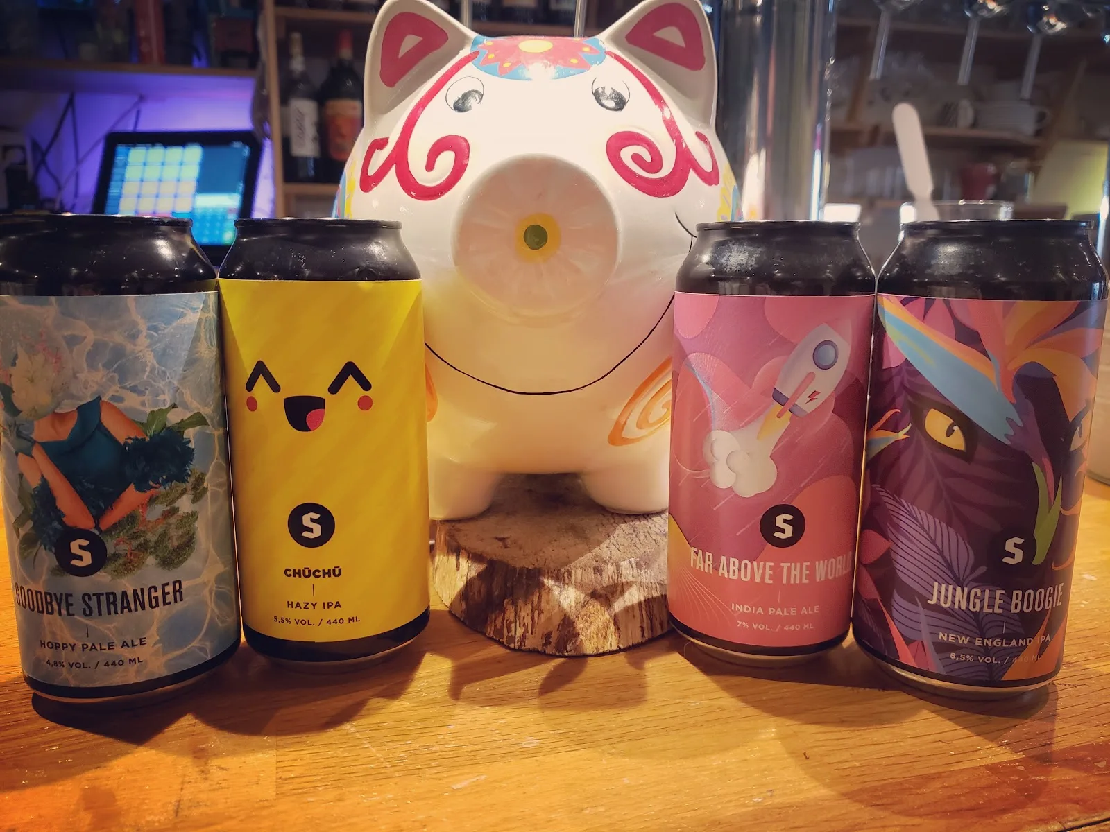 Quatre canettes de bière artisanale alignées sur le comptoir en bois de L'Instant T, devant une tirelire cochon en céramique